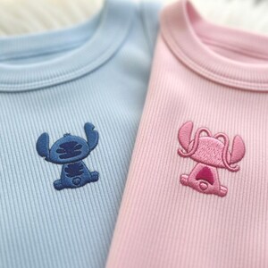 May include: Two ribbed knit bodysuits, one light blue and one pink. Each features an embroidered cartoon character on the chest. The blue bodysuit has a blue character, and the pink one has a pink character.
