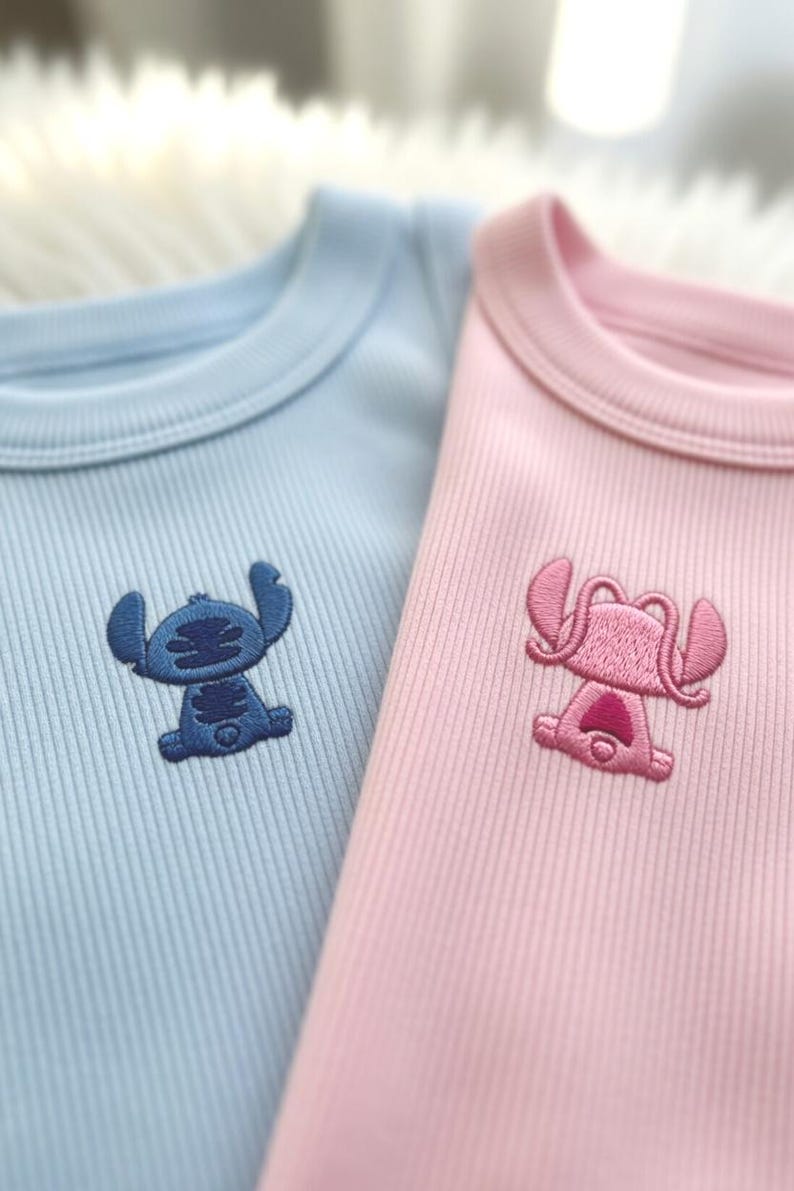 May include: Two ribbed knit bodysuits, one light blue and one pink. Each features an embroidered cartoon character on the chest. The blue bodysuit has a blue character, and the pink one has a pink character.