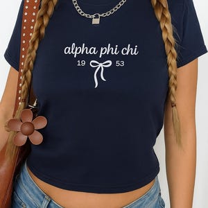 Puede incluir: Top corto azul marino con el texto "alpha phi chi" sobre los números "19" y "53" y un lazo. El top se combina con vaqueros azules. También se ve un bolso de cuero marrón con un adorno en forma de flor.