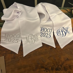 May include: Four white satin graduation stoles with embroidered text and monograms. Two stoles have "HOCO 2025" in silver and blue, and two have custom monograms in silver and blue thread. The stoles are displayed on a wooden surface.