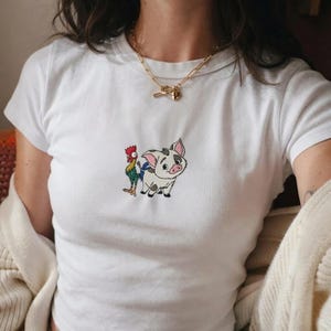 Może przedstawiać: Biały t-shirt z kr&oacute;tkim rękawem z małym, wyśrodkowanym haftem przedstawiającym świnkę i koguta z kresk&oacute;wki. Świnka jest biała w czarne łaty z r&oacute;żowymi uszami, a kogut ma wielokolorowe czerwone, zielone i ż&oacute;łte pi&oacute;ra.