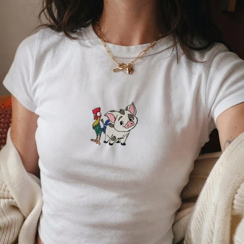 Może przedstawiać: Biały t-shirt z kr&oacute;tkim rękawem z małym, wyśrodkowanym haftem przedstawiającym świnkę i koguta z kresk&oacute;wki. Świnka jest biała w czarne łaty z r&oacute;żowymi uszami, a kogut ma wielokolorowe czerwone, zielone i ż&oacute;łte pi&oacute;ra.