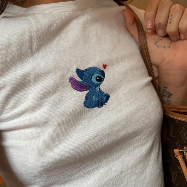 May include: White t-shirt featuring an embroidered blue cartoon character with purple ears and a small red heart. The character has large eyes and is sitting down. The shirt is made of a soft, white fabric.