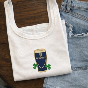 Op de afbeelding: Witte tanktop met een Guinness pint-ontwerp. Het ontwerp toont een marineblauw pintglas met een gouden rand en het Guinness-logo, samen met twee groene klavers. De tanktop is gemaakt van een geribbelde stof.