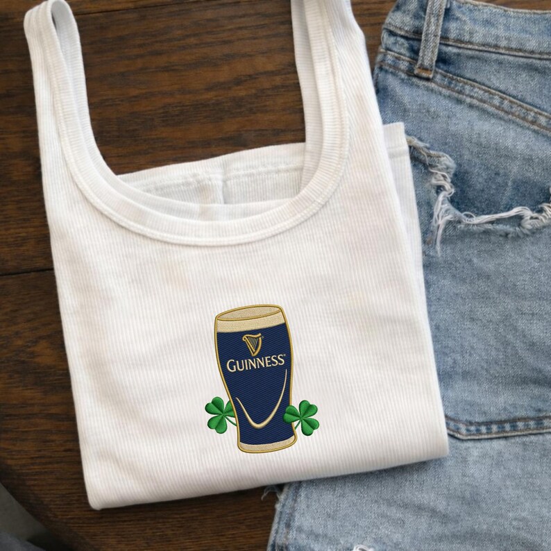 Op de afbeelding: Witte tanktop met een Guinness pint-ontwerp. Het ontwerp toont een marineblauw pintglas met een gouden rand en het Guinness-logo, samen met twee groene klavers. De tanktop is gemaakt van een geribbelde stof.