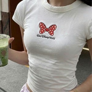 K&ouml;nnte beinhalten: Wei&szlig;es T-Shirt mit einer roten Schleife mit wei&szlig;en Punkten und dem Text "Our Disney World". Das kurz&auml;rmelige Shirt hat einen Rundhalsausschnitt. Die Schleife ist rot umrandet und hat wei&szlig;e Punkte. Das Shirt ist aus weichem Material.