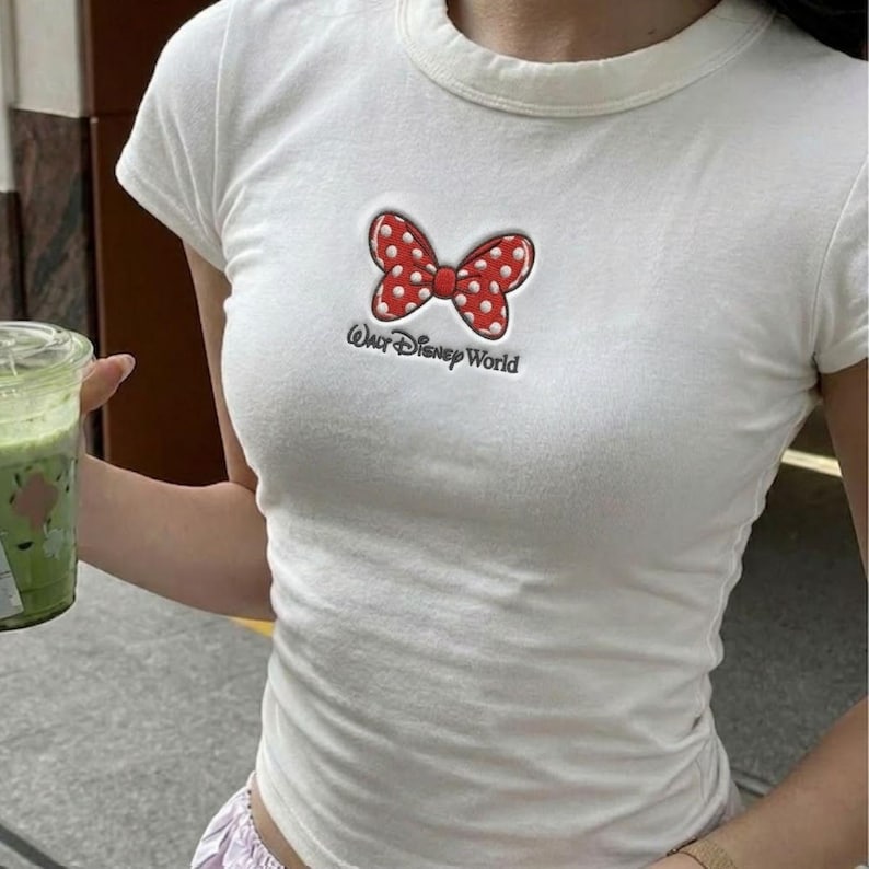 K&ouml;nnte beinhalten: Wei&szlig;es T-Shirt mit einer roten Schleife mit wei&szlig;en Punkten und dem Text "Our Disney World". Das kurz&auml;rmelige Shirt hat einen Rundhalsausschnitt. Die Schleife ist rot umrandet und hat wei&szlig;e Punkte. Das Shirt ist aus weichem Material.