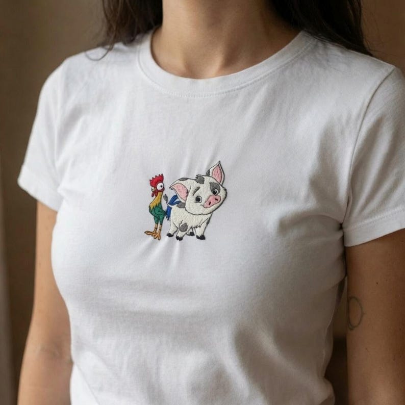 Może przedstawiać: Biały t-shirt z kr&oacute;tkim rękawem z małym, wyśrodkowanym haftem przedstawiającym rysunkową świnkę w czarne łaty oraz rysunkowego koguta z czerwonym grzebieniem, zielonymi pi&oacute;rami i ż&oacute;łtymi łapami, umieszczonym na wysokości klatki piersiowej.