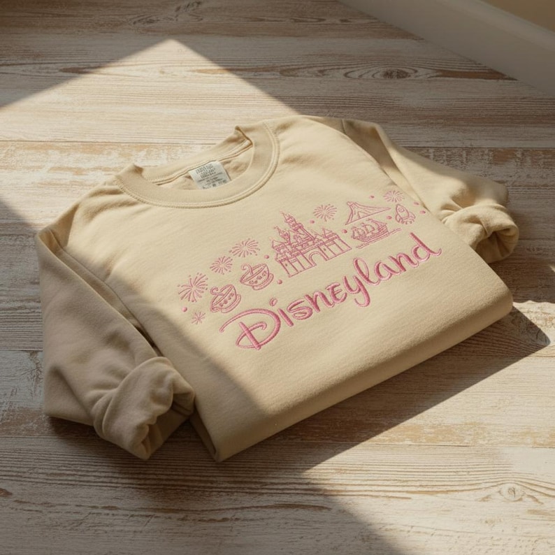 May include: A beige crewneck sweatshirt with the word "Disneyland" embroidered in pink script. Above the word are pink embroidered images of a castle, fireworks, and other park icons. The sweatshirt is folded on a wooden surface.