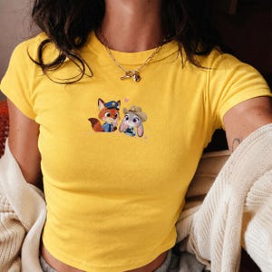 May include: Yellow crop top featuring a cartoon fox and bunny in police and sheriff uniforms, with pink heart accents. The shirt has short sleeves and a crew neck. A gold necklace is visible.