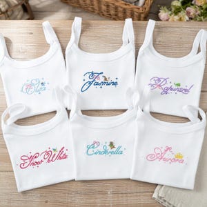 May include: Six white tank tops with embroidered names: Elsa, Jasmine, Rapunzel, Snow White, Cinderella, and Aurora. Each top features a different color of script and small decorative elements, such as crowns and butterflies.
