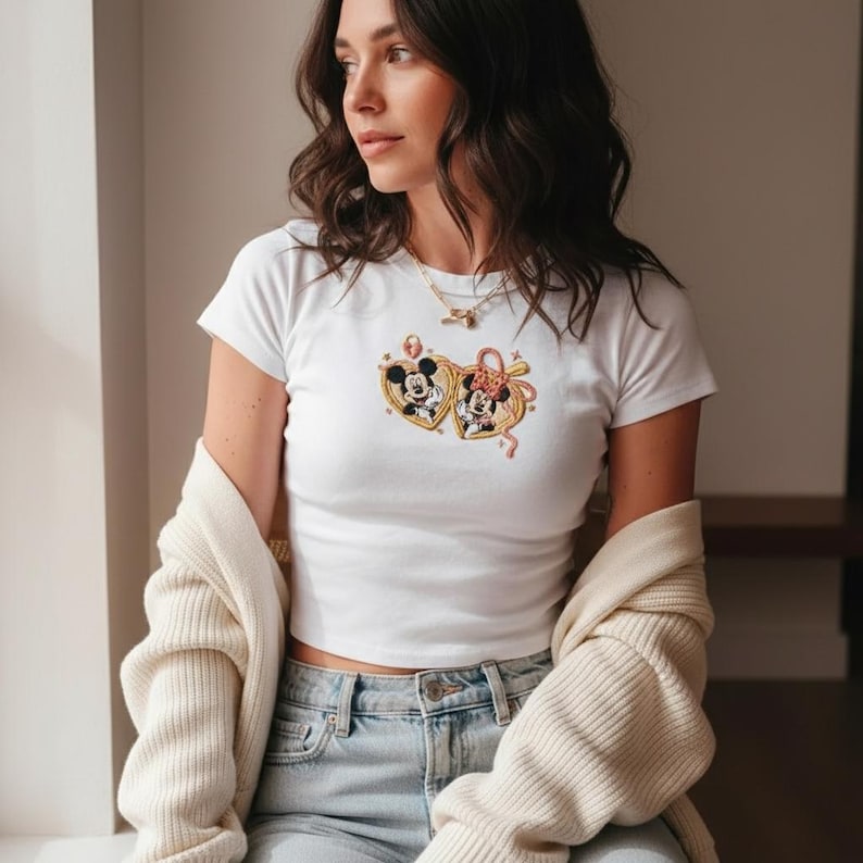 Puede incluir: Camiseta blanca con un dise&ntilde;o de Mickey y Minnie Mouse dentro de marcos en forma de coraz&oacute;n. El dise&ntilde;o incluye detalles dorados y rosas. La camiseta de manga corta se combina con vaqueros de lavado claro y una rebeca crema.