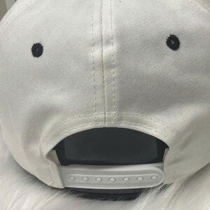 May include: White baseball cap with a black button on top and two black eyelets. The adjustable strap is white. The cap is made of a textured fabric.