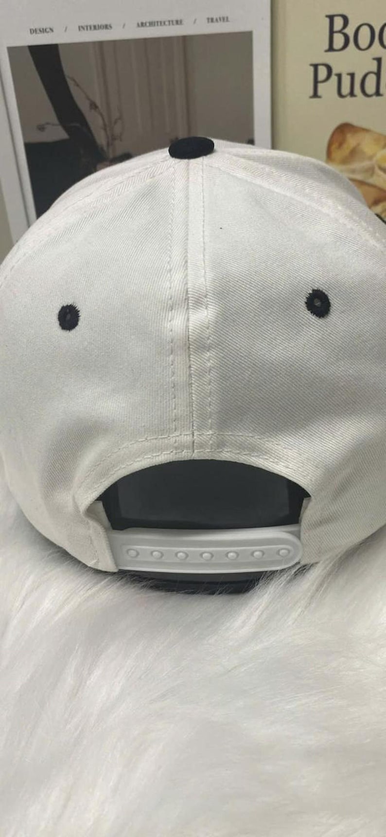 May include: White baseball cap with a black button on top and two black eyelets. The adjustable strap is white. The cap is made of a textured fabric.