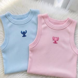 May include: Two ribbed tank tops, one light blue and one pink, each with an embroidered cartoon character. The blue tank top features a blue character, while the pink tank top has a pink character. The tops are displayed on a fluffy white surface.