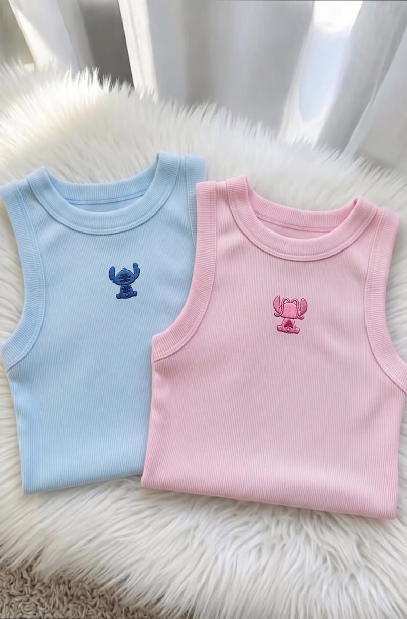 May include: Two ribbed tank tops, one light blue and one pink, each with an embroidered cartoon character. The blue tank top features a blue character, while the pink tank top has a pink character. The tops are displayed on a fluffy white surface.