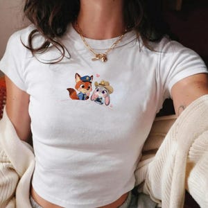 May include: White t-shirt featuring a cartoon fox and bunny in police and cowboy attire, with pink heart accents. The shirt has short sleeves and a crew neck.