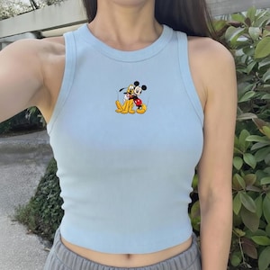 May include: Light blue ribbed tank top featuring a Mickey Mouse and Pluto graphic. The sleeveless top has a round neckline and is made of a soft, ribbed material. The graphic shows Mickey Mouse and Pluto hugging each other.