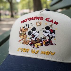 May include: A baseball cap with a cream-colored crown and a navy blue brim. The cap features an embroidered design of Mickey and Minnie Mouse, Pluto, and the text "NOTHING CAN STOP US NOW" in red and yellow.