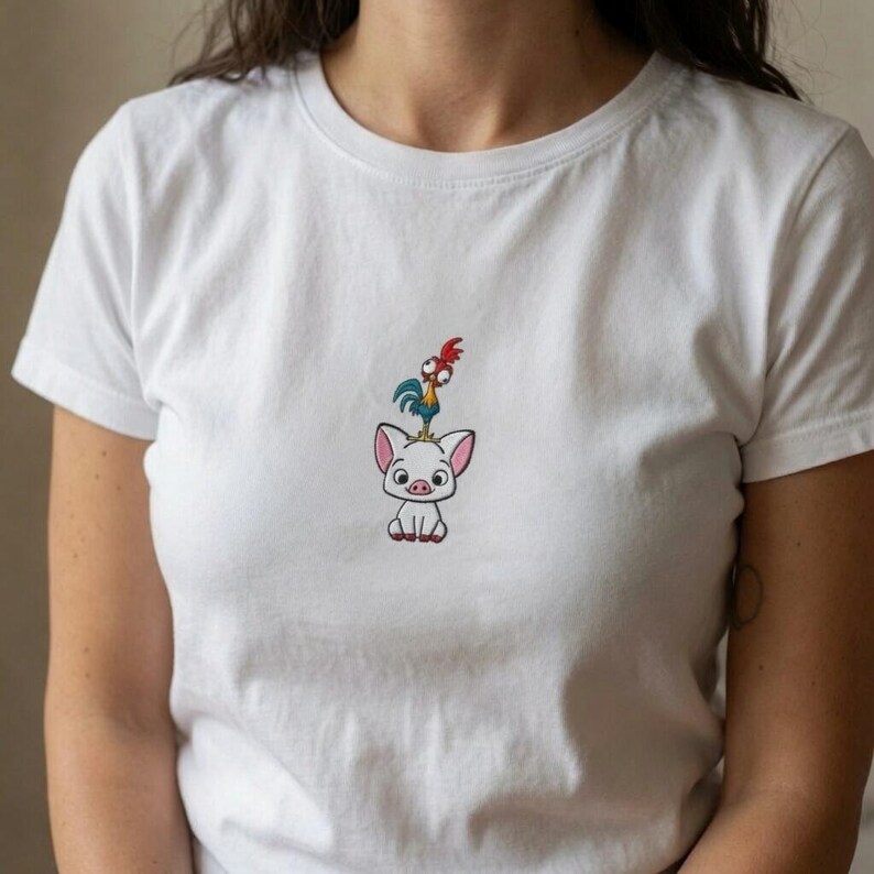 May include: A white short-sleeved t-shirt featuring a small, centered embroidery of a cartoon pig with a small rooster standing on its head. The pig is white with pink ears and a pink snout, while the rooster has blue feathers and a red comb.