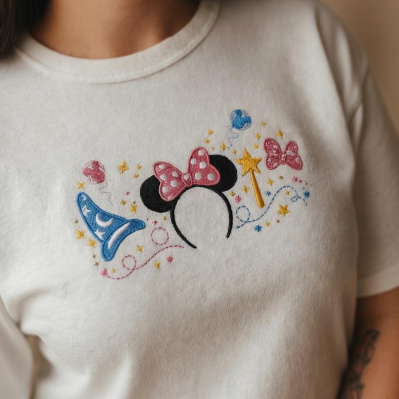 May include: A white t-shirt featuring an embroidered design of black mouse ears with a pink polka-dot bow, a blue wizard hat with a white moon, a yellow star wand, pink and blue balloons, and small yellow stars scattered across the chest area.