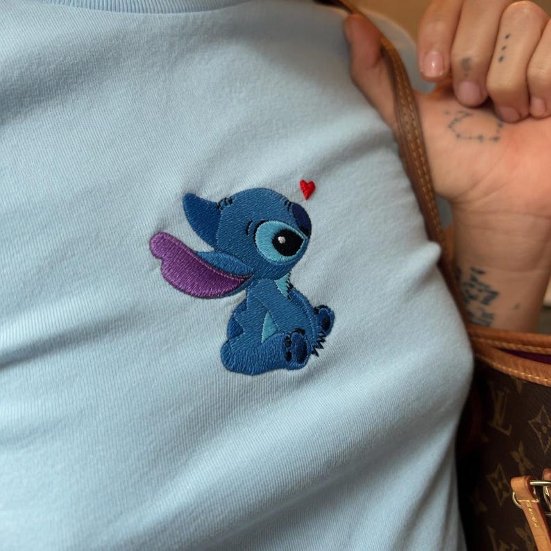 May include: Light blue t-shirt featuring an embroidered Stitch character with a red heart above its head. The character is in shades of blue and purple. The t-shirt is made of a soft material.