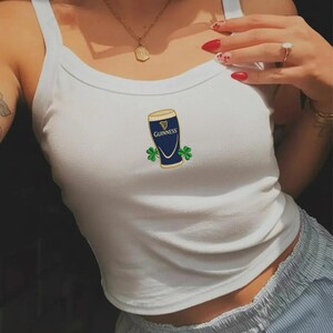 May include: White tank top with a Guinness beer glass graphic. The graphic features a blue glass with the Guinness logo and two green shamrocks. The tank top is a casual style.