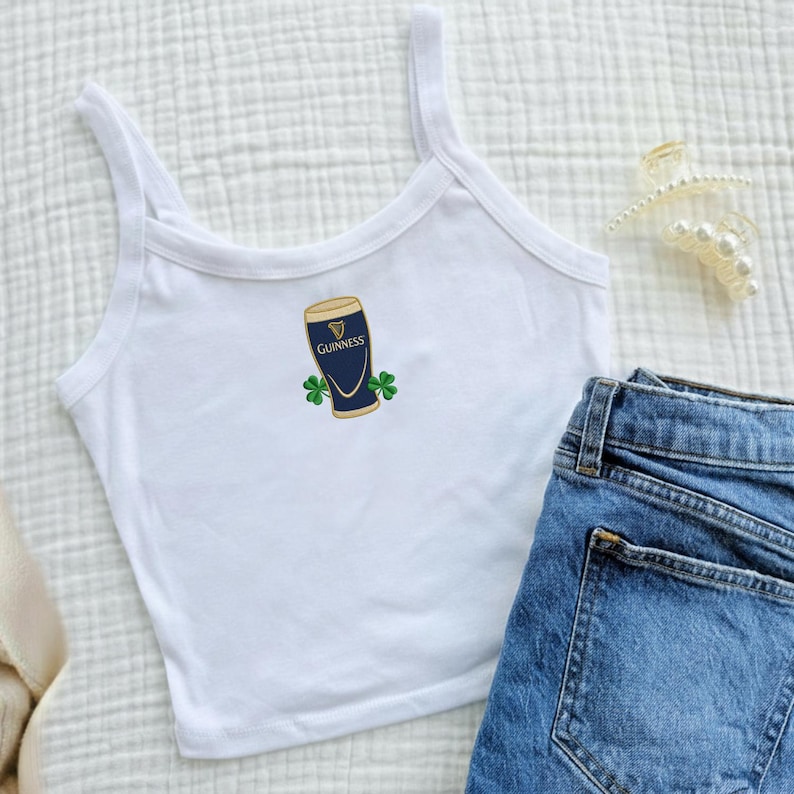 May include: White tank top with a Guinness pint graphic and shamrocks. The tank top is paired with blue denim jeans. A pearl hair clip is visible in the background.