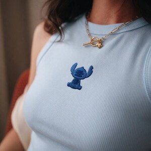 May include: Light blue ribbed tank top with a dark blue embroidered Stitch character. A gold-colored chain necklace with a heart pendant is worn with the top. The top is a solid light blue color.