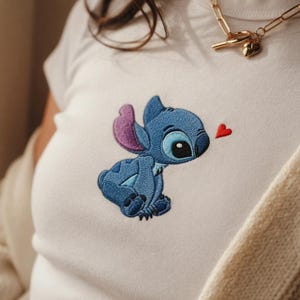 May include: White t-shirt featuring an embroidered image of the character Stitch from the Disney movie Lilo & Stitch. The blue Stitch is looking at a small red heart. The shirt has a round neckline.