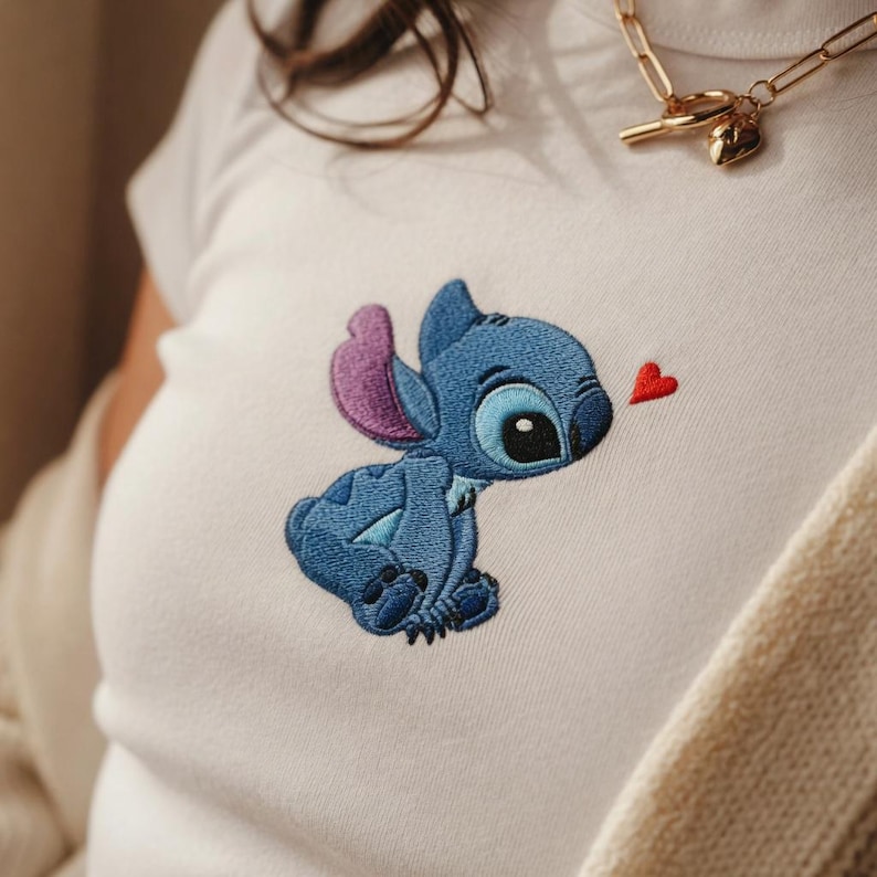 May include: White t-shirt featuring an embroidered image of the character Stitch from the Disney movie Lilo & Stitch. The blue Stitch is looking at a small red heart. The shirt has a round neckline.