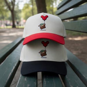 May include: Two baseball caps stacked on top of each other. The top cap has a white crown and red brim, with an embroidered image of a cartoon character holding a Mickey Mouse balloon. The bottom cap has a white crown and a black brim.