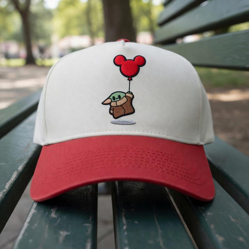 May include: A baseball cap with a white crown and a red brim. The cap features an embroidered design of a green character holding onto a red Mickey Mouse balloon. The cap is sitting on a green bench.