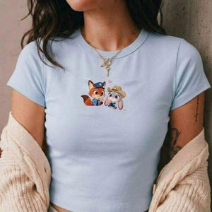 May include: Light blue t-shirt featuring a cartoon fox in a police uniform and a bunny wearing a cowboy hat. The graphic includes small pink hearts. The shirt has a crew neck and short sleeves.