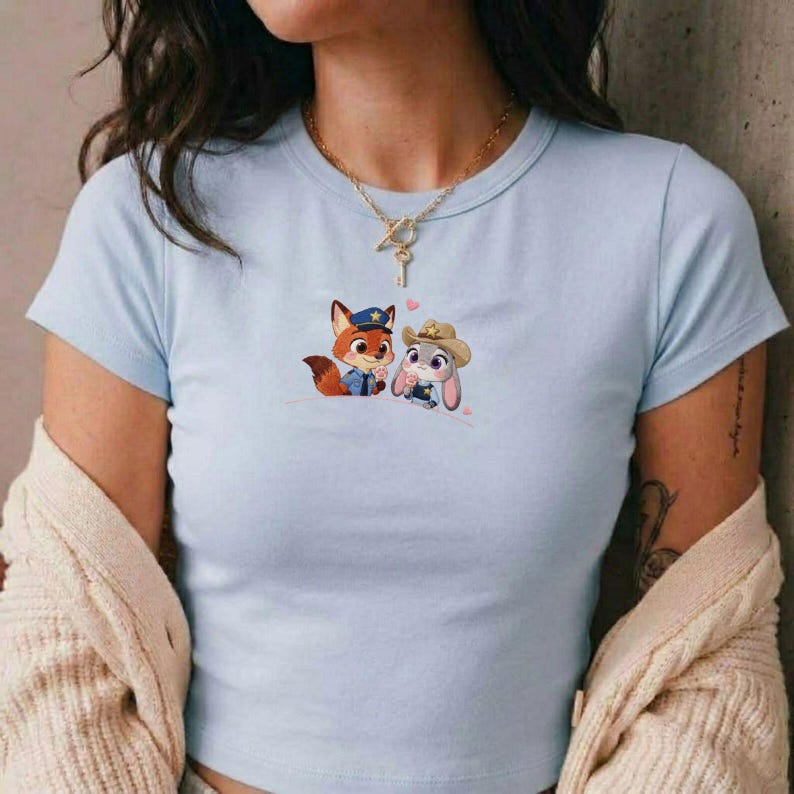 May include: Light blue t-shirt featuring a cartoon fox in a police uniform and a bunny wearing a cowboy hat. The graphic includes small pink hearts. The shirt has a crew neck and short sleeves.