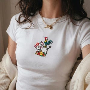 K&ouml;nnte beinhalten: Ein wei&szlig;es Kurzarm-T-Shirt mit einem zentralen gestickten Aufn&auml;her, der ein kleines Schwein mit einer roten Blume auf dem Kopf und einen Hahn auf seinem R&uuml;cken zeigt, umgeben von bunten Blumen. Das T-Shirt wird mit einer Goldkette kombiniert.