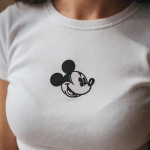 May include: White t-shirt featuring an embroidered Mickey Mouse design. The iconic cartoon character is depicted in black, white, and light gray, with a smiling expression. The shirt has a crew neck and short sleeves, suitable for casual wear.