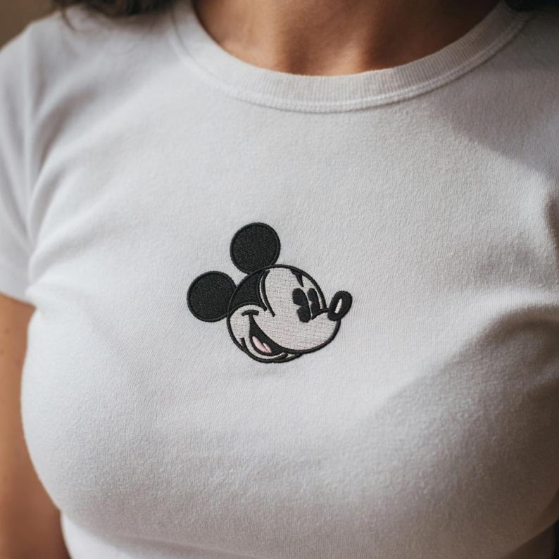 May include: White t-shirt featuring an embroidered Mickey Mouse design. The iconic cartoon character is depicted in black, white, and light gray, with a smiling expression. The shirt has a crew neck and short sleeves, suitable for casual wear.