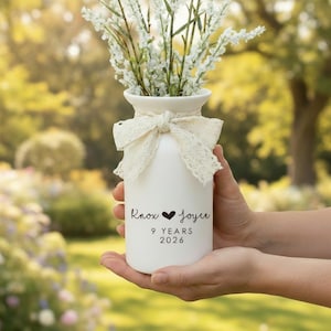 Könnte beinhalten: Eine weiße Keramikvase mit einer Spitzenschleife und weißen Blumen. Die Vase trägt die Namen "Knox" und "Joyce" mit einem Herzsymbol und dem Text "9 YEARS 2026".