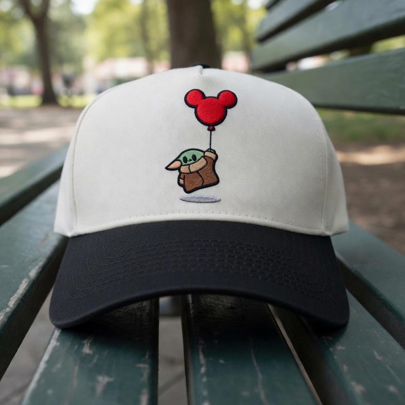 May include: A baseball cap with a white crown and a black brim. The cap features an embroidered design of a green character holding a red Mickey Mouse balloon. The cap is likely made of cotton or a similar fabric.