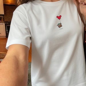 Peut inclure: T-shirt blanc avec une illustration de dessin anim&eacute; d'une petite cr&eacute;ature verte tenant un ballon rouge en forme de c&oelig;ur. Le motif est centr&eacute; sur la poitrine. Le t-shirt a des manches courtes et un col rond.