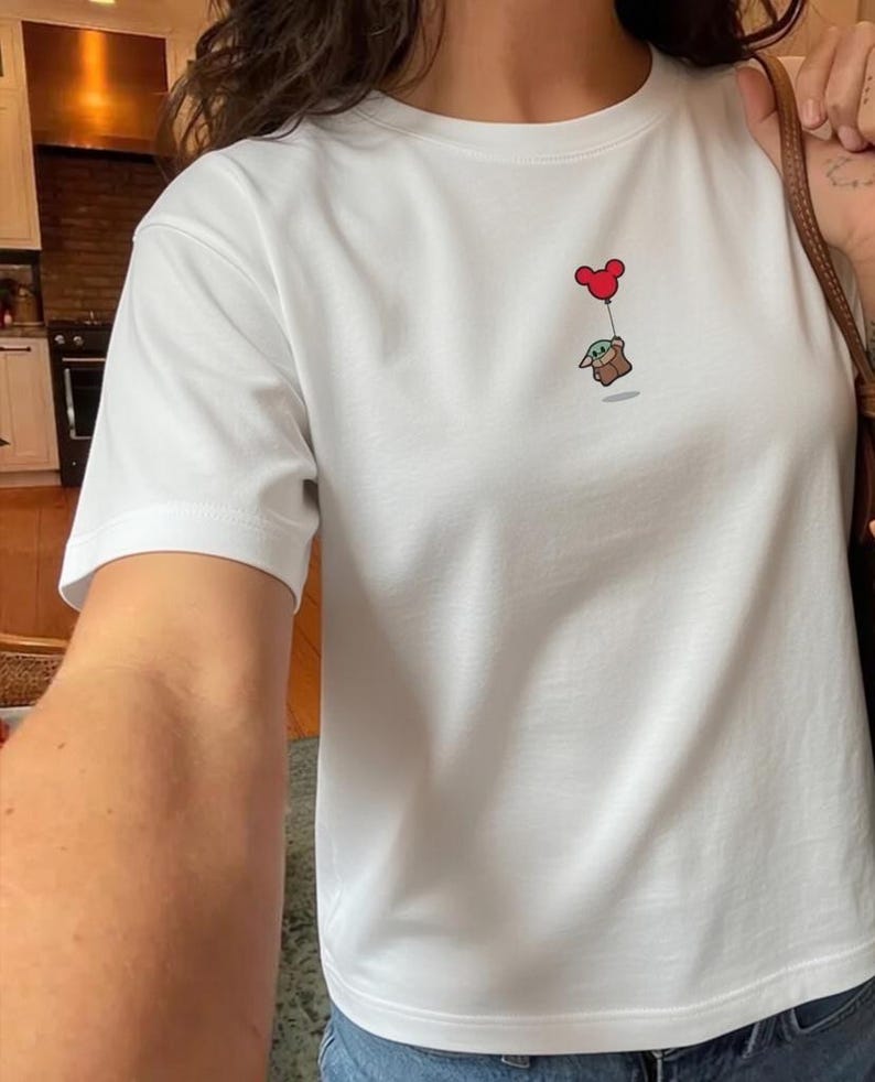 Peut inclure: T-shirt blanc avec une illustration de dessin anim&eacute; d'une petite cr&eacute;ature verte tenant un ballon rouge en forme de c&oelig;ur. Le motif est centr&eacute; sur la poitrine. Le t-shirt a des manches courtes et un col rond.