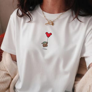 Peut inclure: T-shirt blanc avec une broderie repr&eacute;sentant Grogu, &eacute;galement connu sous le nom de Baby Yoda, tenant un ballon rouge Mickey Mouse. Un collier en or est visible en haut de l'image. Le motif est sur le devant du t-shirt.