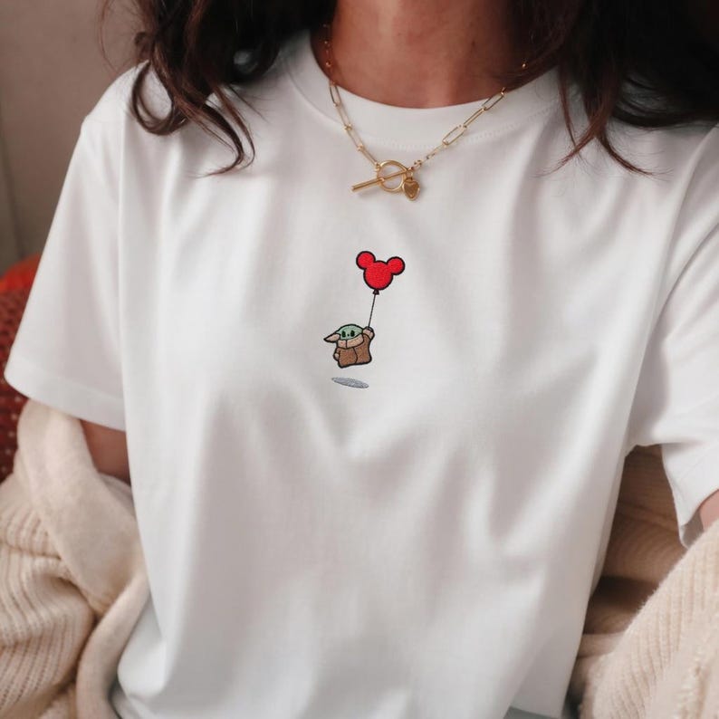 Peut inclure: T-shirt blanc avec une broderie repr&eacute;sentant Grogu, &eacute;galement connu sous le nom de Baby Yoda, tenant un ballon rouge Mickey Mouse. Un collier en or est visible en haut de l'image. Le motif est sur le devant du t-shirt.