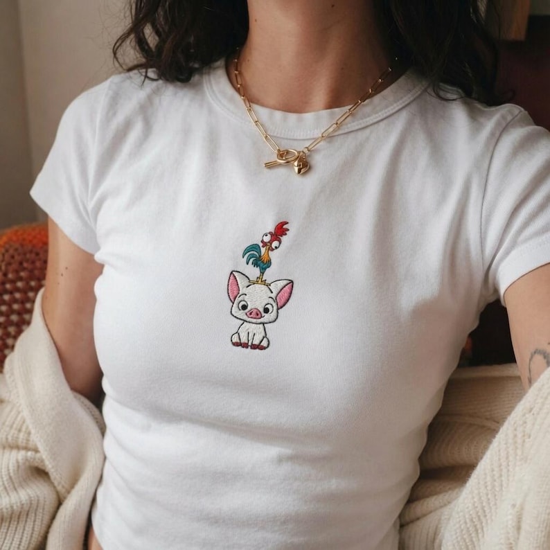 May include: A white short-sleeved t-shirt featuring a small, centered embroidery of a cartoon pig with a rooster standing on its head. The pig is white with pink ears and the rooster has blue, green, and red feathers. A gold chain necklace is worn.