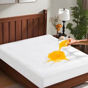 May include: A white mattress protector fitted over a bed, featuring a textured surface. A clear glass is pouring bright orange liquid onto the center of the protector, demonstrating its liquid-resistant properties on the fabric surface.