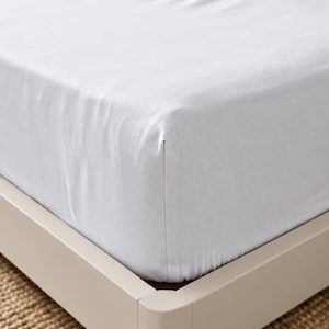 May include: A crisp white fitted sheet pulled taut over the corner of a mattress. The fabric has a smooth, clean texture and is neatly tucked around the edge of the bed frame, showing a simple, solid color design with no patterns or embellishments.