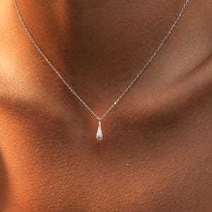 Puede incluir: Un delicado collar de plata con un pequeño colgante en forma de lágrima. El colgante cuelga de una fina cadena, creando un aspecto minimalista y elegante. El collar se muestra sobre un fondo de tono de piel.