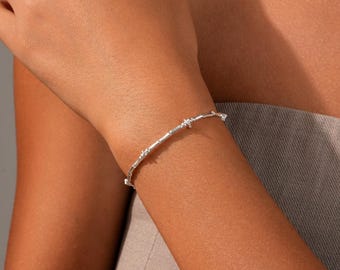 Könnte beinhalten: Ein dünnes, poliertes Armband in Silberoptik mit einer Reihe kleiner, gleichmäßig verteilter Knoten entlang des Bandes. Das metallische Accessoire hat eine glatte, reflektierende Oberfläche und eine feine Struktur, die das Handgelenk umschließt.