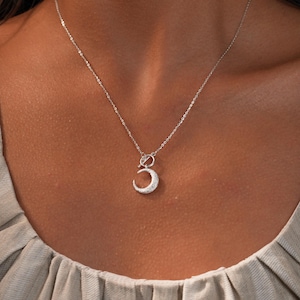 May include: A silver necklace featuring a crescent moon pendant. The moon is encrusted with small, sparkling stones. The necklace is delicate and hangs on a thin chain, creating a minimalist and elegant look.